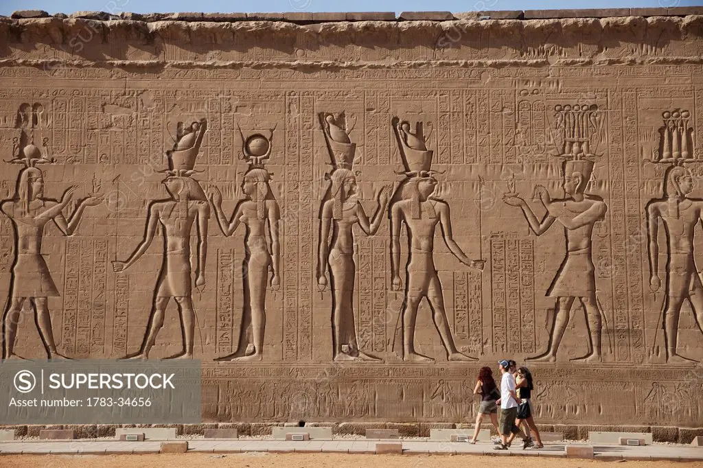 Tourists walking past carvings of egyptian gods and pharoahs rear wall of edfu temple; edfu upper egypt