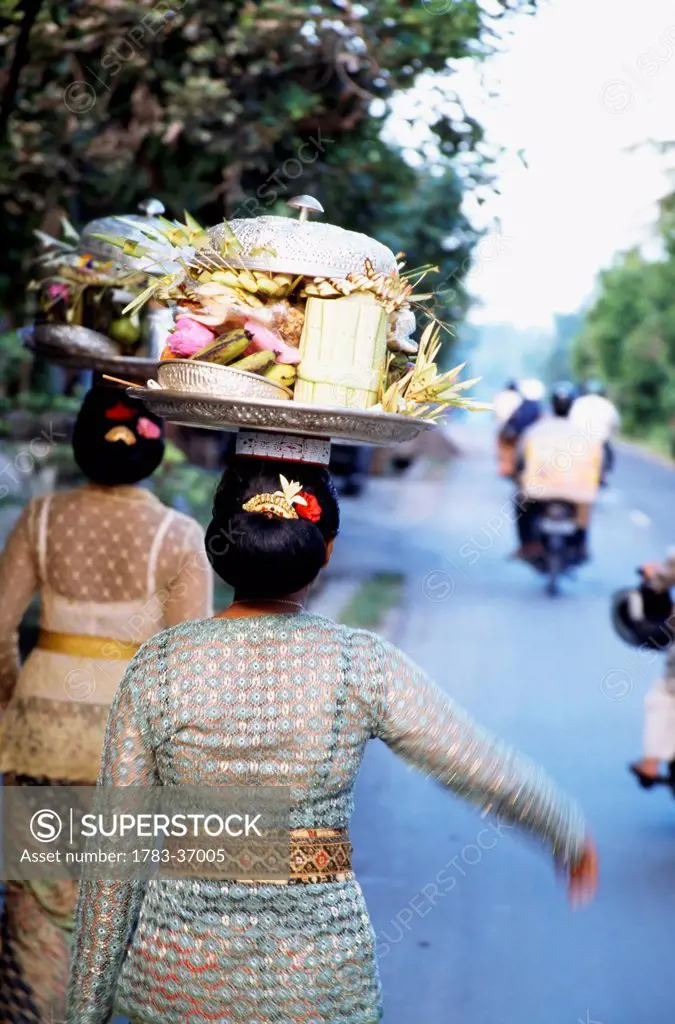 Woman ( Women ) In Embroidered Dress With Temple Offerings Walking Down A Road With Motorcyclists In The Distance . Ubud,Bali,Indonesia .