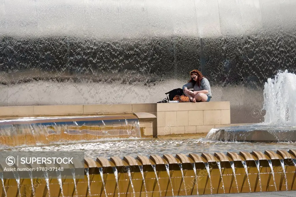 Woman reading book beside fountains outside Sheffield train station; South Yorkshire, UK