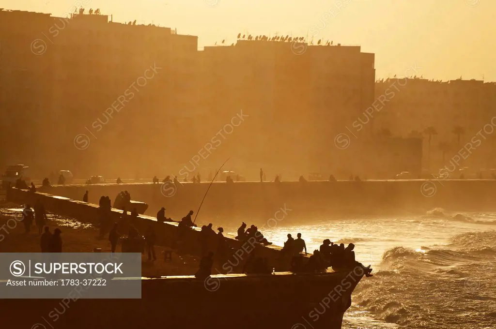 People sitting and fishing at dusk from sea wall beside Hassan II Mosque; Casablanca, Morocco