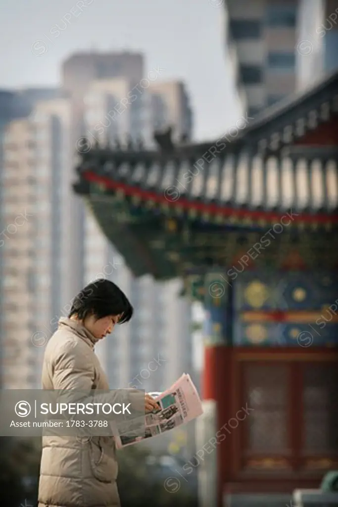 A local woman reads a newspaper, in the background there is a  contrast of old and new, a traditional Chinese structure beside modern skyscrapers, Beijing, China