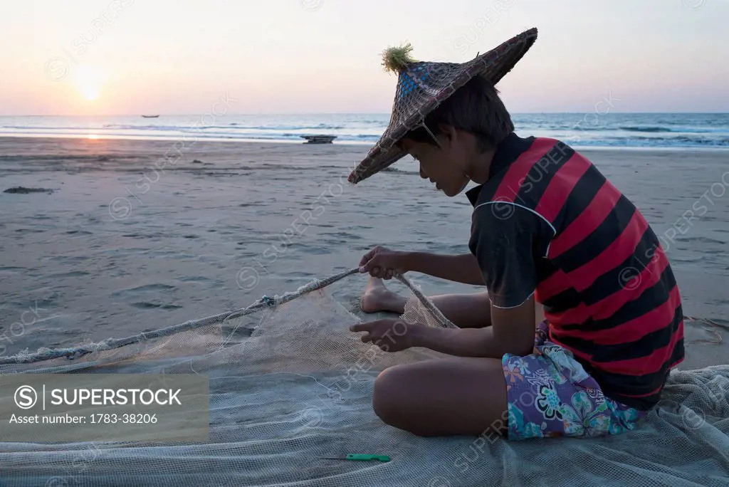 Young man fixing fishing net on beach at sunset; Yea Thoe, Irrawaddyi Division, Myanmar (Burma)