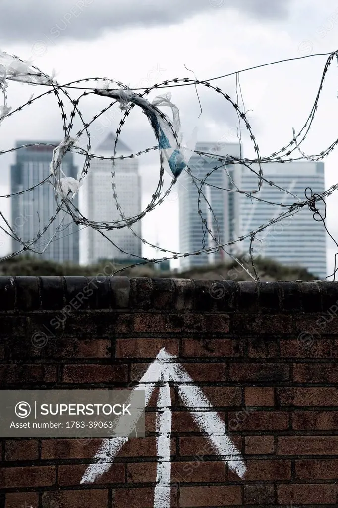 Brick wall with barbed wire on top, plastic bags caught on barbs, Canary Wharf in background; Greenwich Peninsula, London, UK