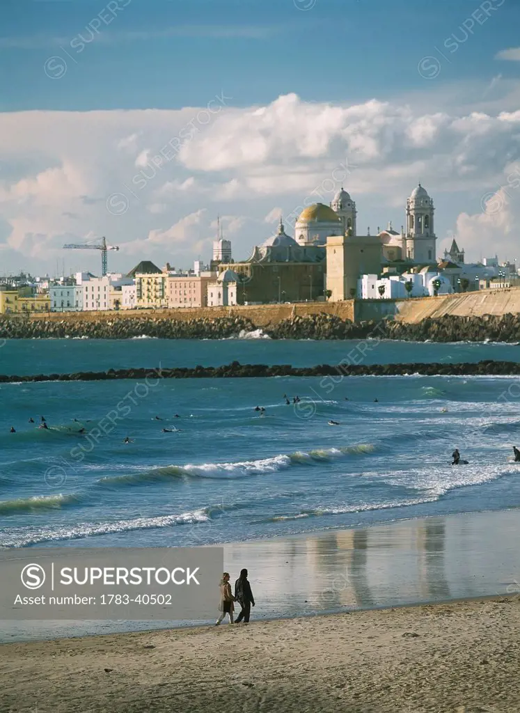 Couple Walking Along Beach In Front Of Surfers On Beach And Santa Cruz Cathedral, Cadiz, Andalucia, Spain.,