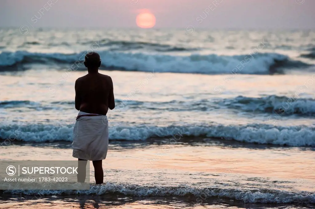 India, Karnataka, Hindu pilgrim praying by ocean at sunset; Gokarna