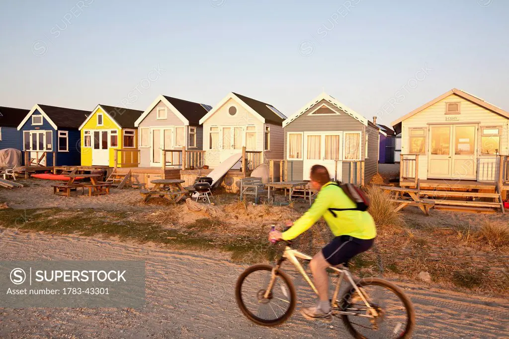 Young man riding mountain bicycle on beach near beach huts; Christchurch Harbour, Mudeford, Dorset, England, United Kingdom