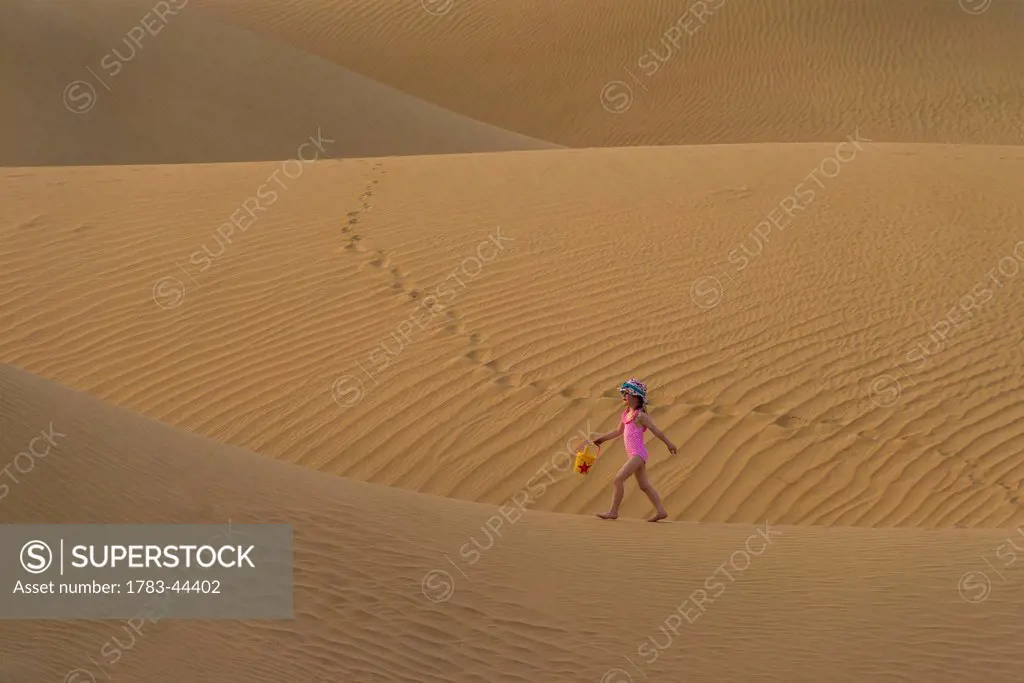Girl in pink swimsuit with bucket and spade walking across sand dunes at dusk; Dubai, United Arab Emirates