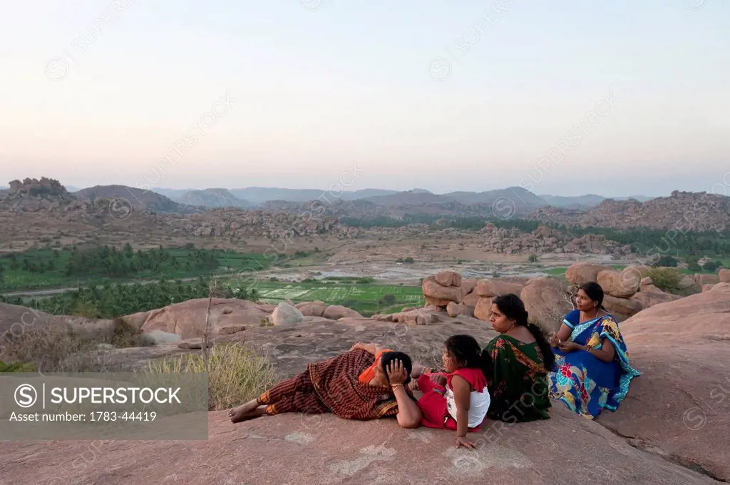 Three women and a girl sit on a rock surface overlooking the landscape; Hampi, Karnataka, India