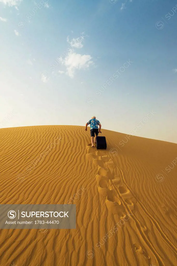 Barefoot man with suitcase walking up sand dune; Dubai, United Arab Emirates