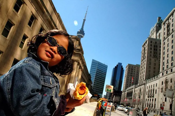 Young girl / child eating hotdog in front of buildings, CN Tower in background, Toronto, Canada