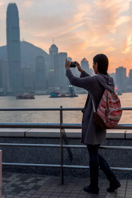 A young woman takes a picture with her camera of the harbour and Hong Kong skyline at sunset, Kowloon; Hong Kong, China
