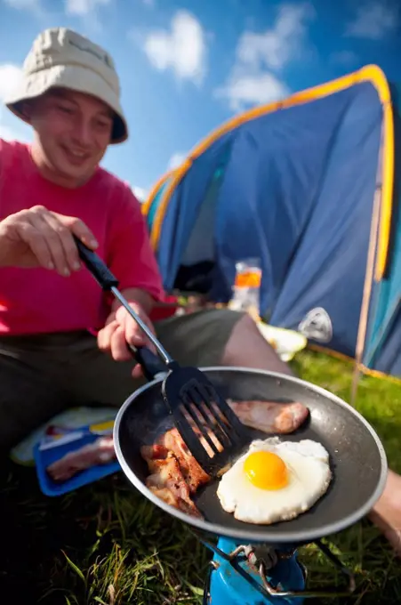 Man cooking fried breakfast of bacon and egg beside tent; Lulworth, Dorset, UK