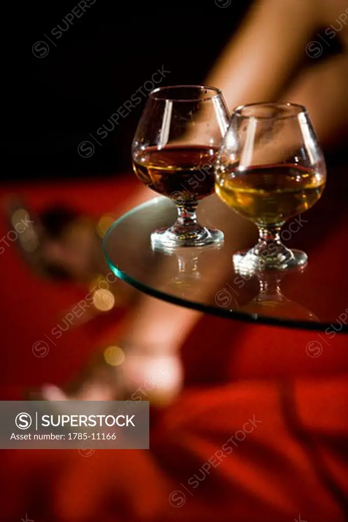 Two brandy glasses on glass table, with woman's legs in background