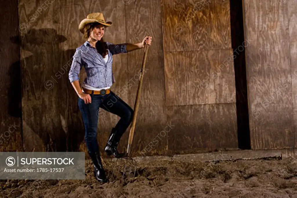 Portrait of young cowgirl standing in horse stable with pitchfork