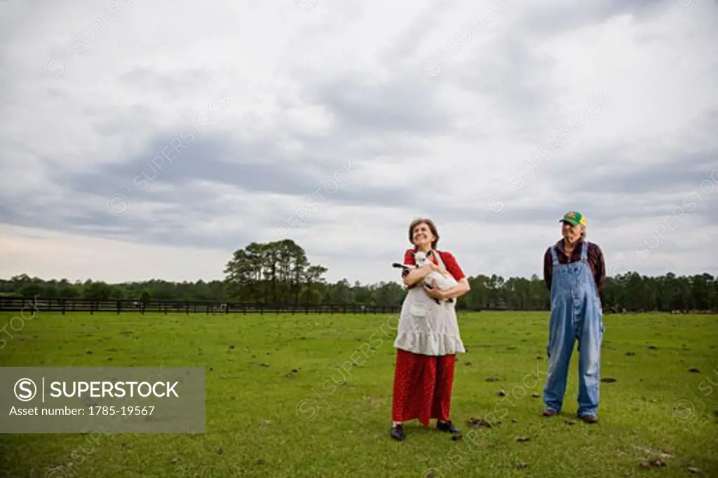 Senior woman holding baby goat out in field, husband in background