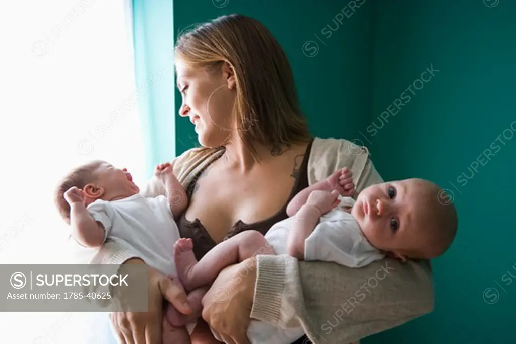 Mother holding newborn twins in arms by window, one crying