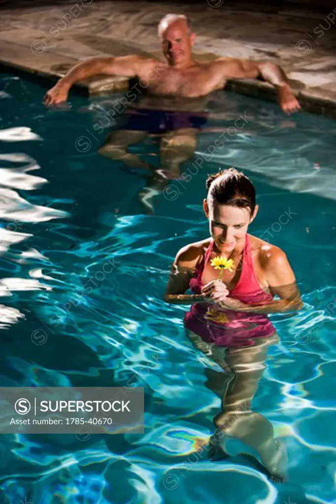 Middle-aged woman holding flower in swimming pool, man in background