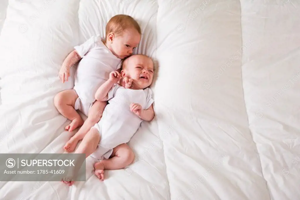 Newborn twin babies lying side by side on soft white blanket, one crying