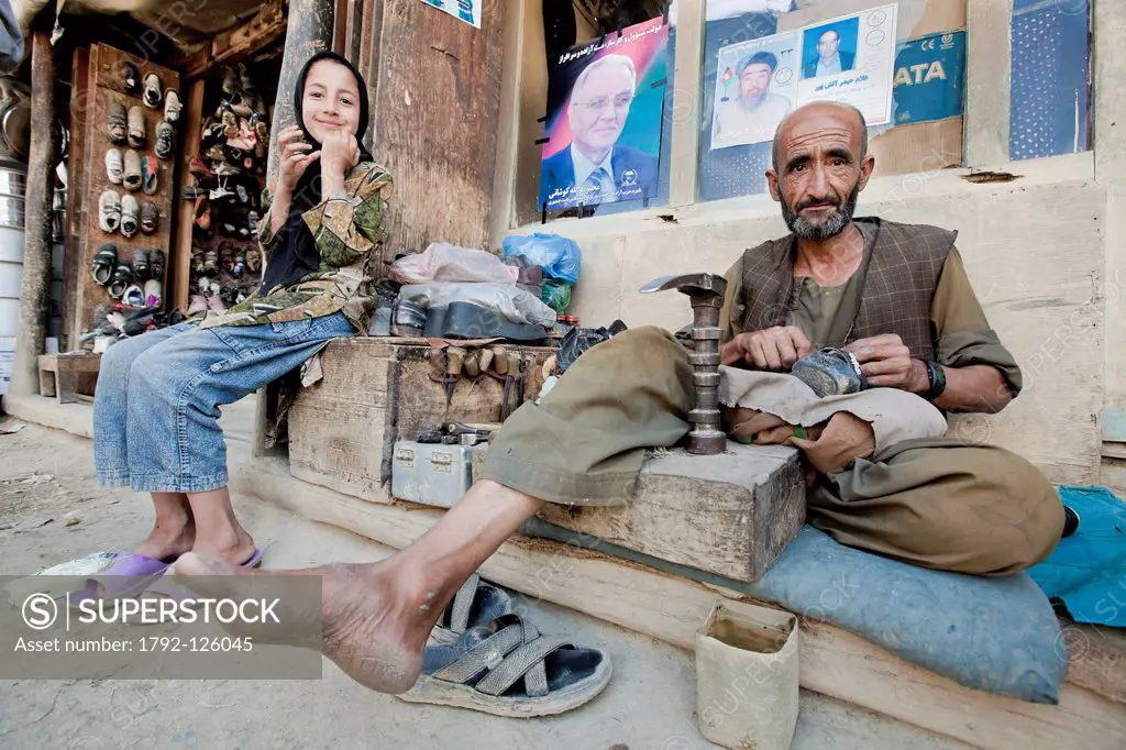 Afghanistan, Badakhshan province, Faizabad, shoemaker and his daughter posing in front of political posters, market, bazaar