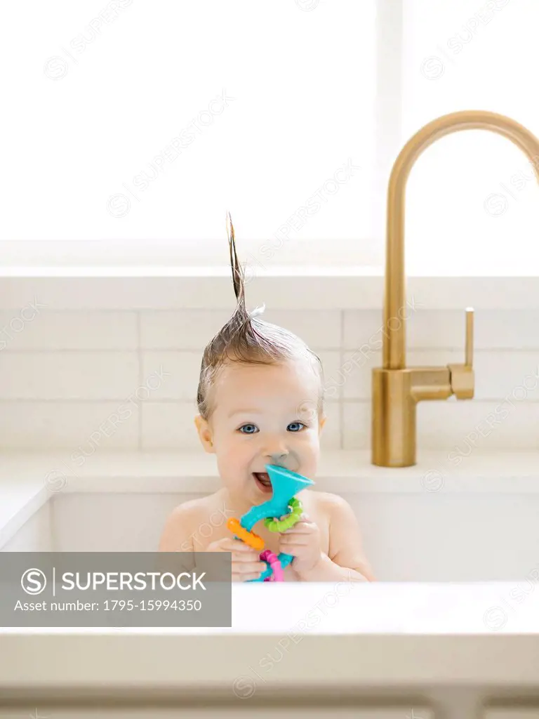 Baby girl with Mohican hairstyle holding toy while bathing in kitchen sink