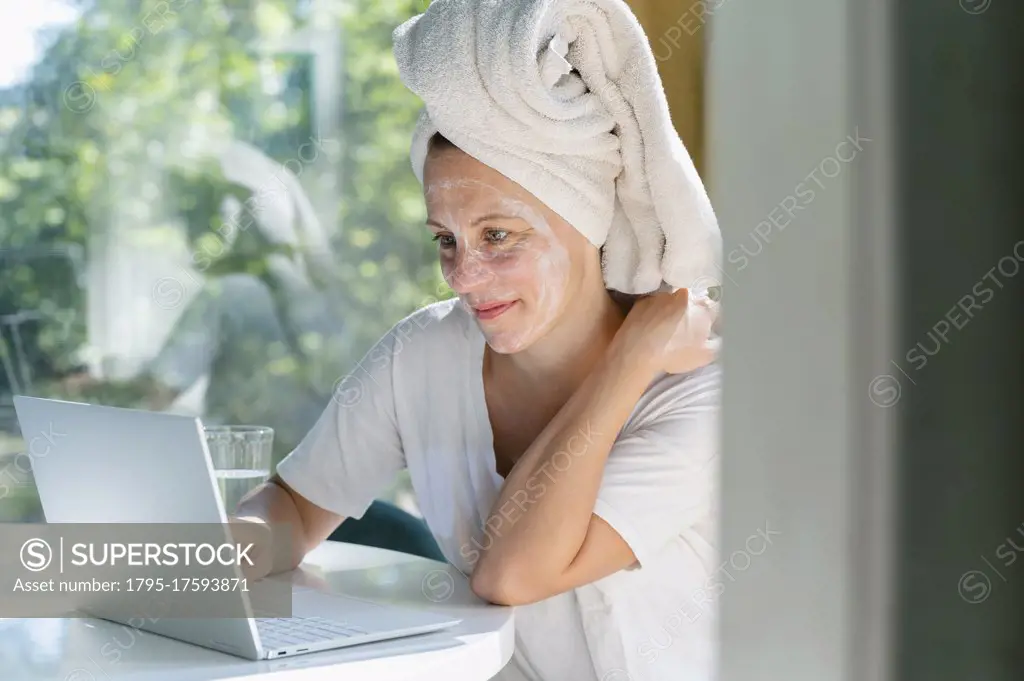 Woman with face mask and towel on head using laptop at table