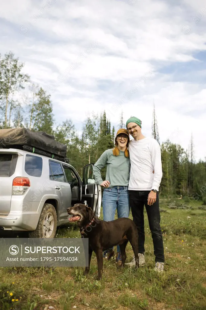 USA, Utah, Uinta National Park, Portrait of smiling couple with dog standing in meadow, off road car in background