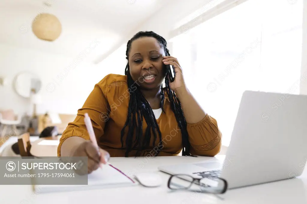 Woman working at desk at home, writing and talking on mobile phone