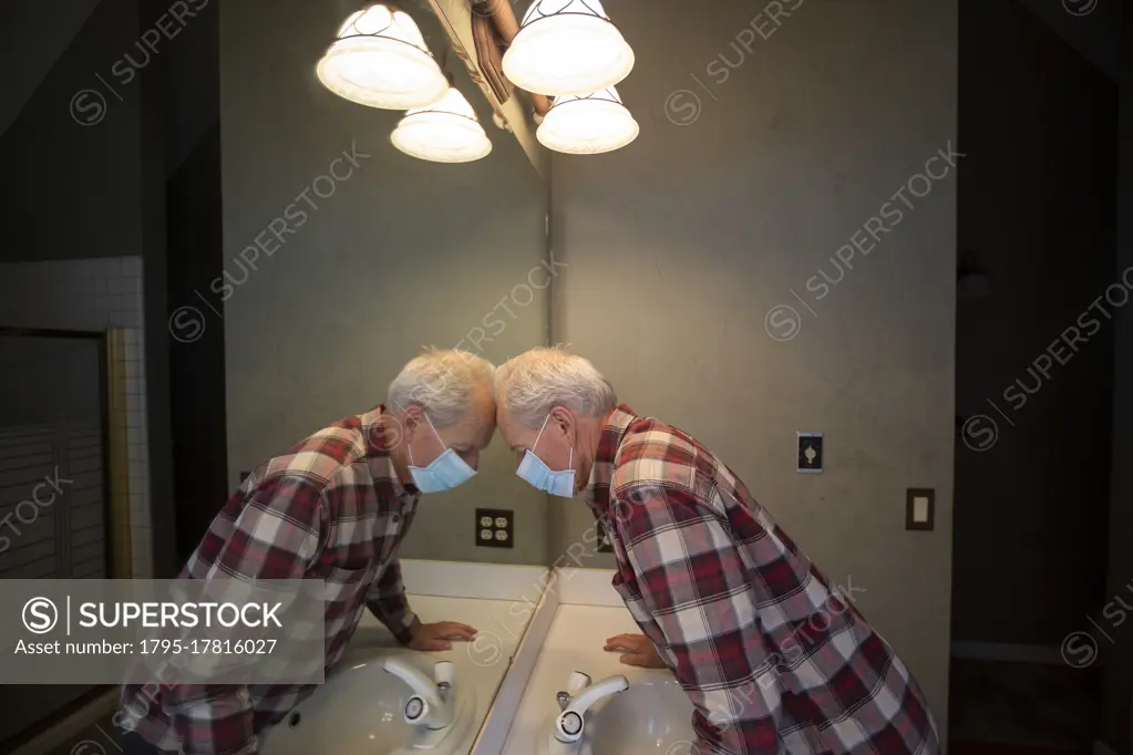 Senior man in Covid protective mask leaning head on mirror in bathroom