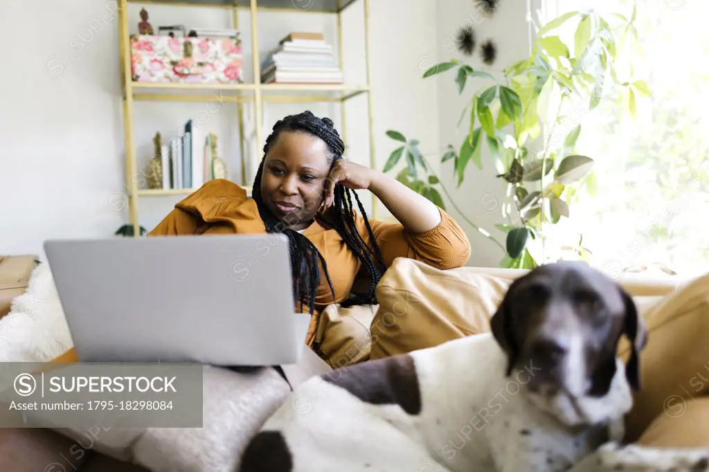 Woman lying on sofa with dog and working on laptop