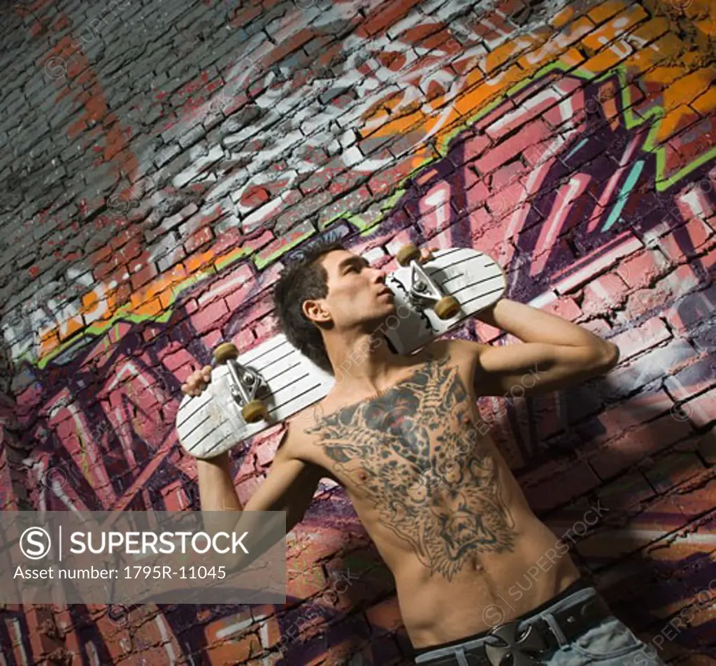 Asian man holding skate board in front of graffitied wall