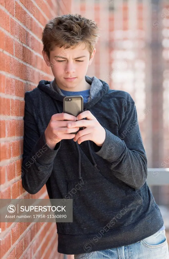Teenage boy (16-17) leaning against brick wall texting