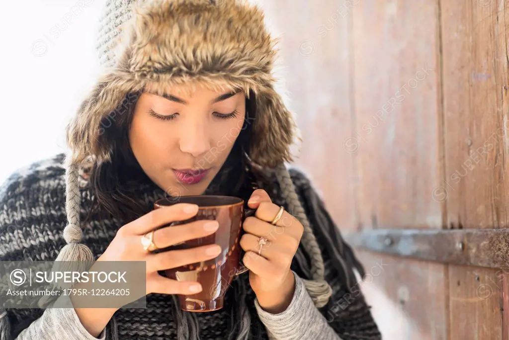Portrait of young woman wearing warm hat, blowing on cup of drink