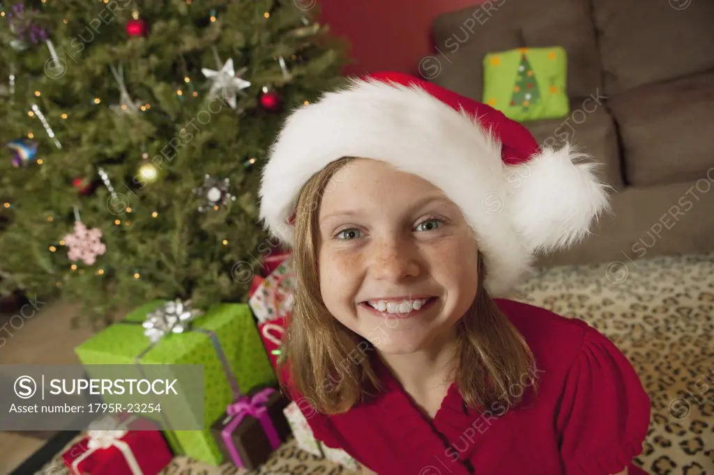 Girl (9) wearing Santa hat sitting in front of Christmas tree, portrait