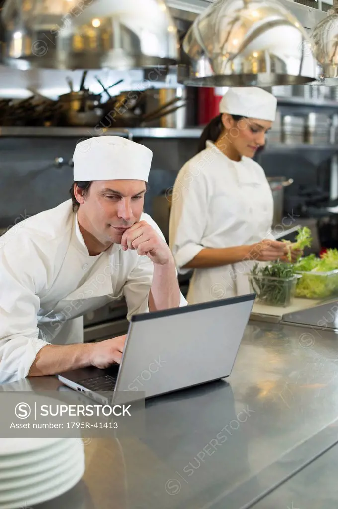 Chef using laptop and cook preparing food in commercial kitchen