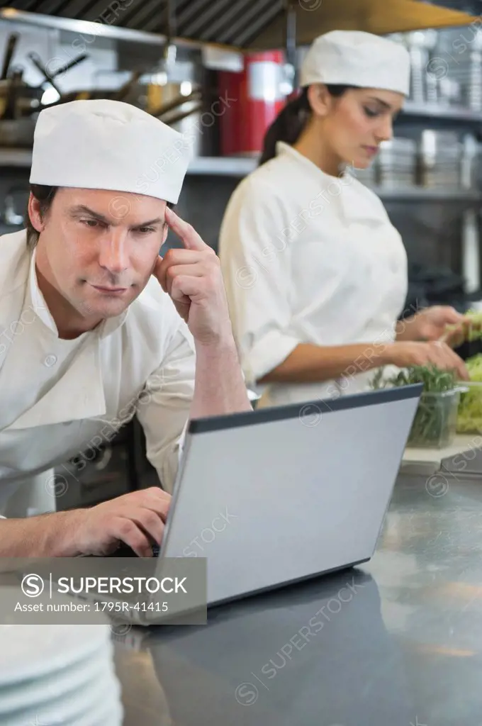 Chef using laptop and cook preparing food in commercial kitchen