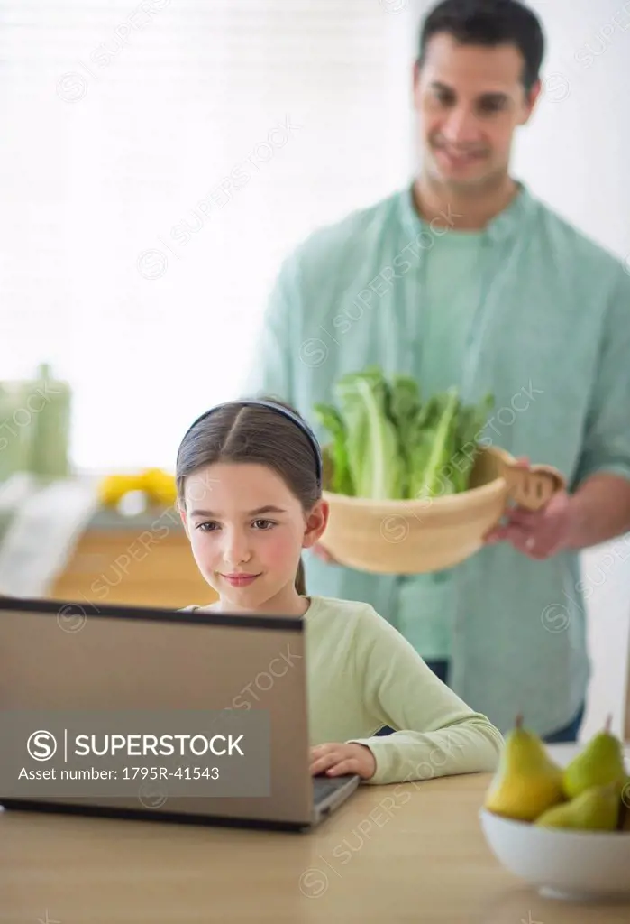 Girl 8_9 using laptop and father preparing food in domestic kitchen