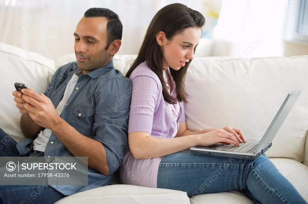Couple sitting on sofa back to back using laptop and phone