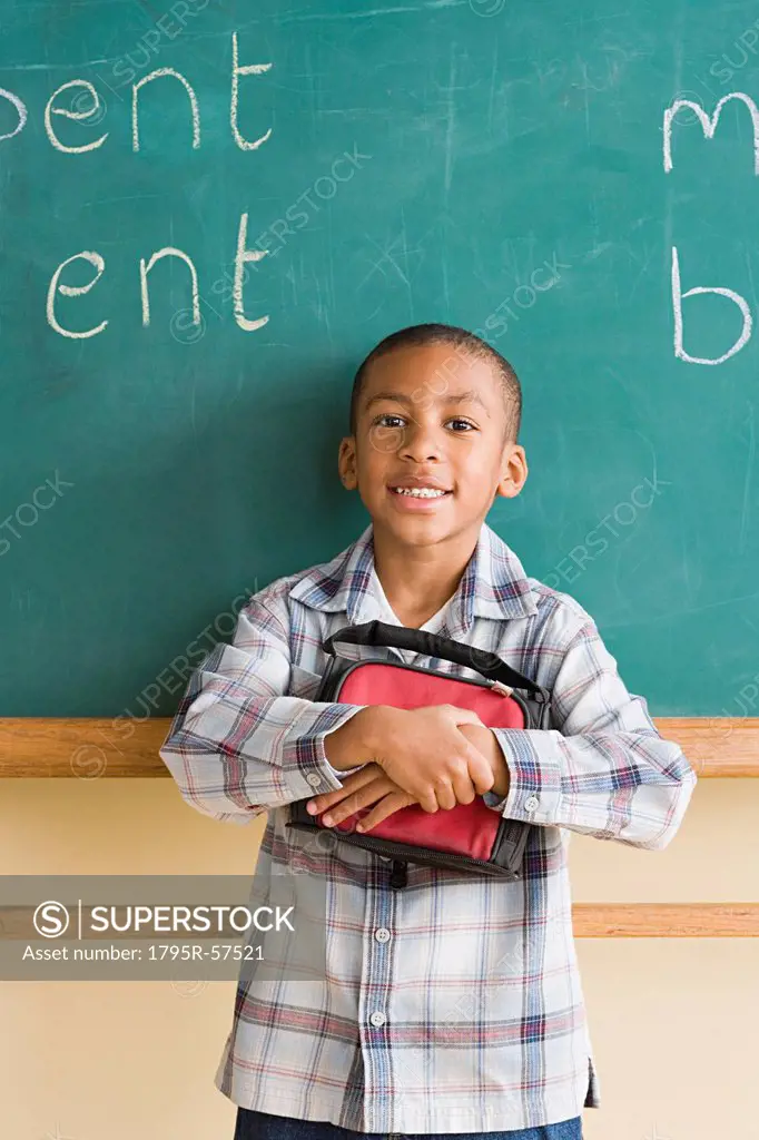 Smiling boy 6_7 holding lunch box against blackboard