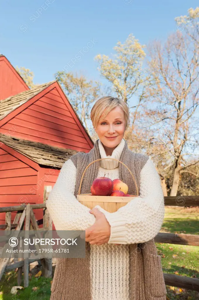 USA, New Jersey, Portrait of smiling woman holding basket with apples in front of cottage house in Autumn