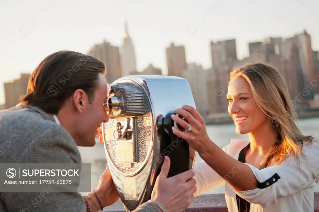 USA, New York, Long Island City, Young couple looking through coin operated binoculars, Manhattan skyline in background