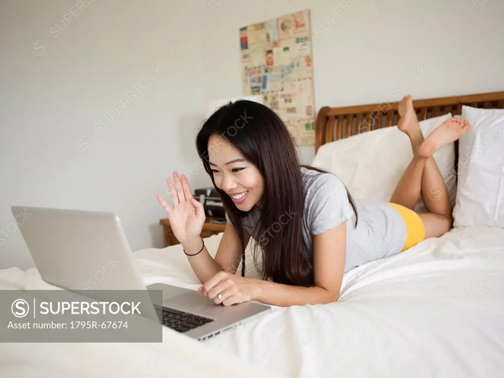 Young woman lying down in bed, using laptop, waving