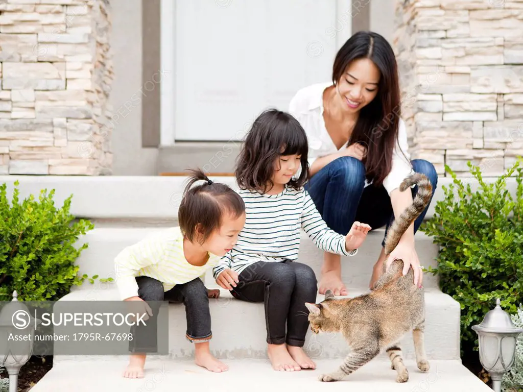 Mother sitting on doorsteps with two daughters 2_3, 4_5 and playing with cat