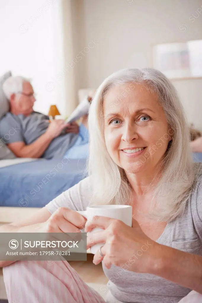 Senior woman holding mug, man sitting on bed in background