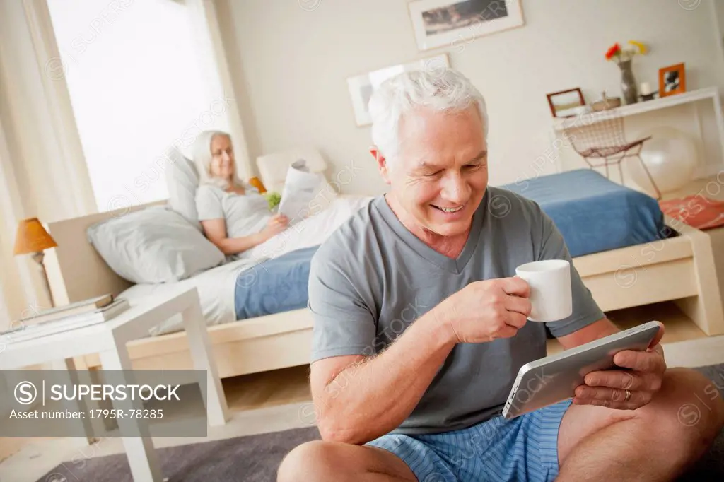 Senior man holding mug, woman sitting on bed in background