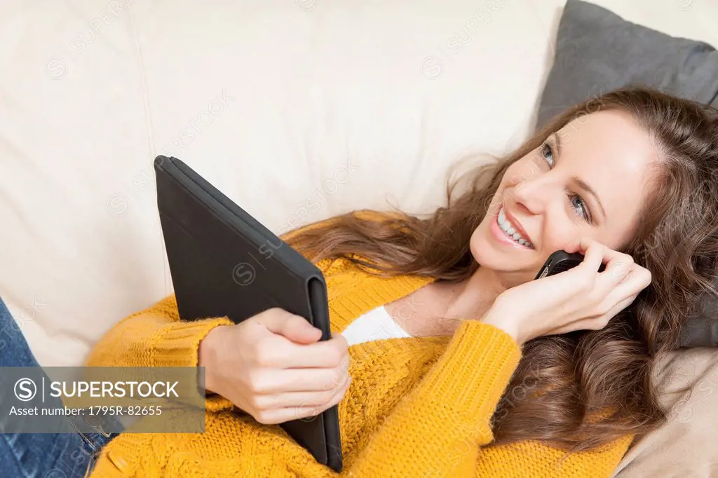 Woman lying on sofa with digital tablet and talking via cell phone