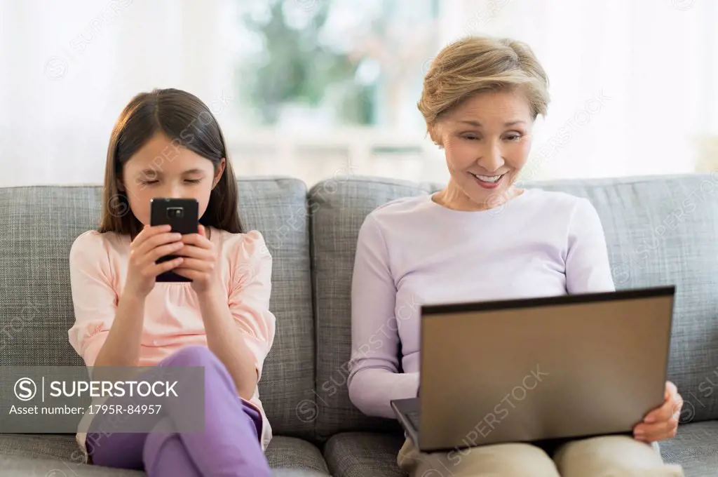 Granddaughter (8-9) and grandmother sitting on sofa and using laptop and cell phone