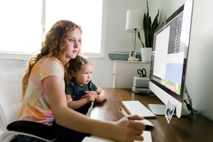 Young mother with her daughter on lap working from home at desk