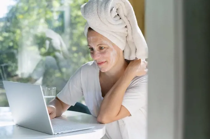 Woman with face mask and towel on head using laptop at table