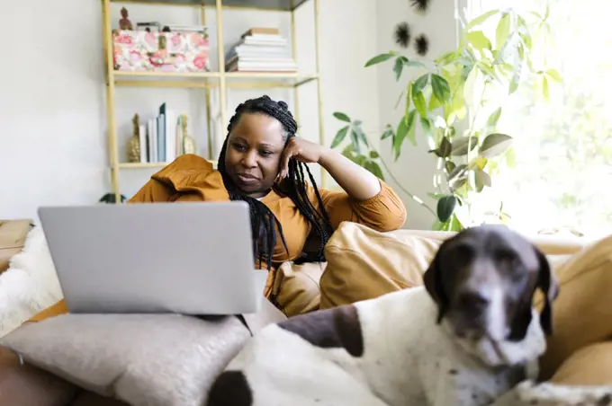 Woman lying on sofa with dog and working on laptop