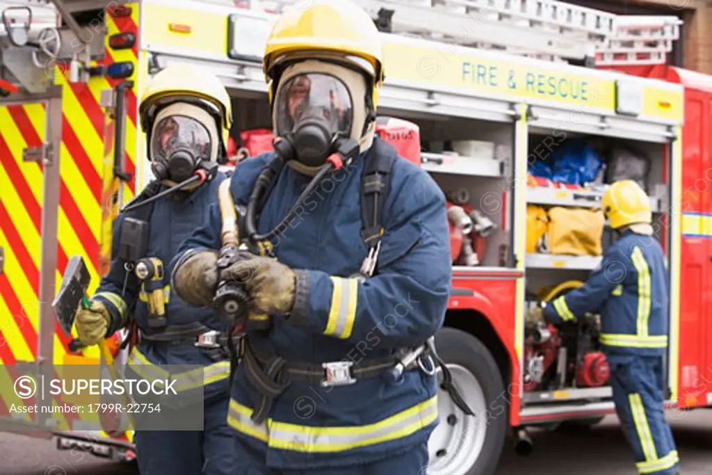 Two firefighters with hose and axe walking away from fire engine and another firefighter in background (selective focus)